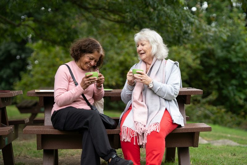Dos amigas mayores conversan y ríen sentadas en una banca del parque